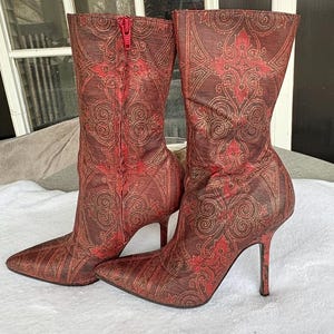 May include: A pair of high-heeled boots with a pointed toe and a red zipper. The boots feature a brown and red ornate pattern with gold accents. The heels are slender and tall.