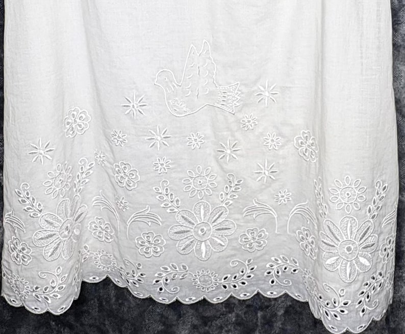 May include: White fabric with intricate embroidered details. The design features a dove, stars, and floral patterns along the hem. The fabric has a scalloped edge, adding a delicate touch to the overall aesthetic.
