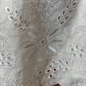 May include: Close-up of white fabric with intricate floral embroidery. The design features large, detailed flowers, smaller blossoms, and delicate leaf patterns. The fabric has a scalloped edge, adding a decorative touch to the overall design.