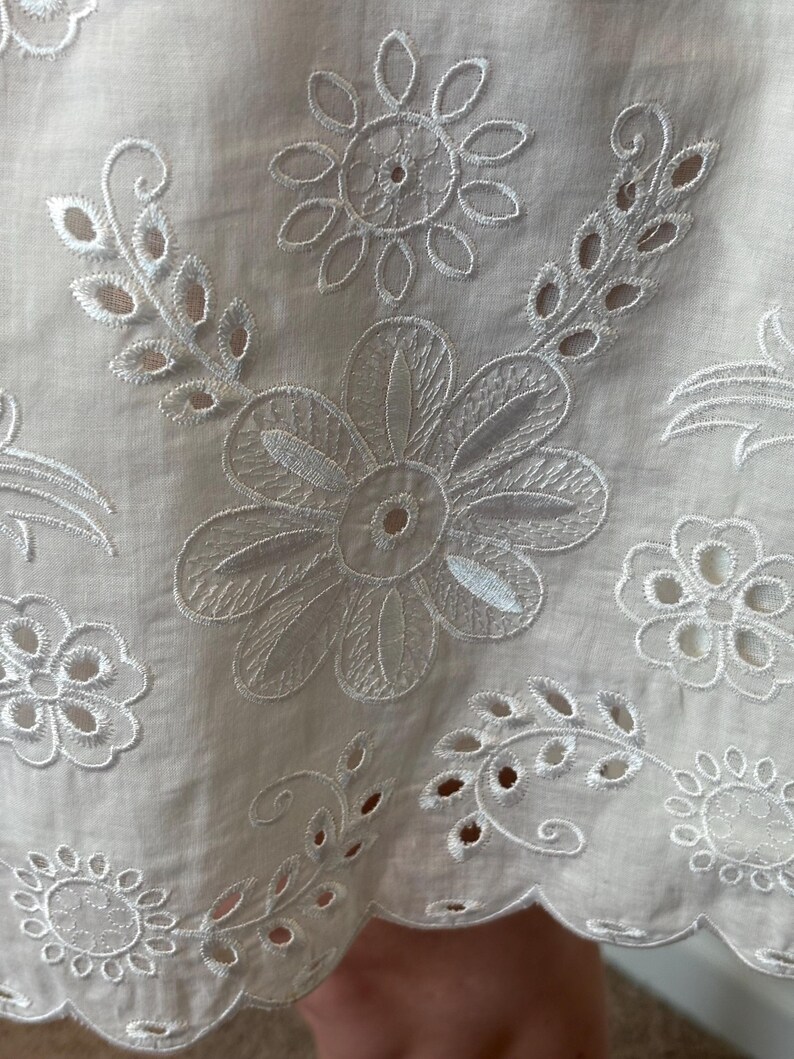 May include: Close-up of white fabric with intricate floral embroidery. The design features large, detailed flowers, smaller blossoms, and delicate leaf patterns. The fabric has a scalloped edge, adding a decorative touch to the overall design.