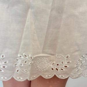 May include: Close-up of a white fabric with a scalloped edge. The edge features an embroidered floral design with cut-out details. The fabric appears to be lightweight and slightly translucent, suggesting a delicate texture.
