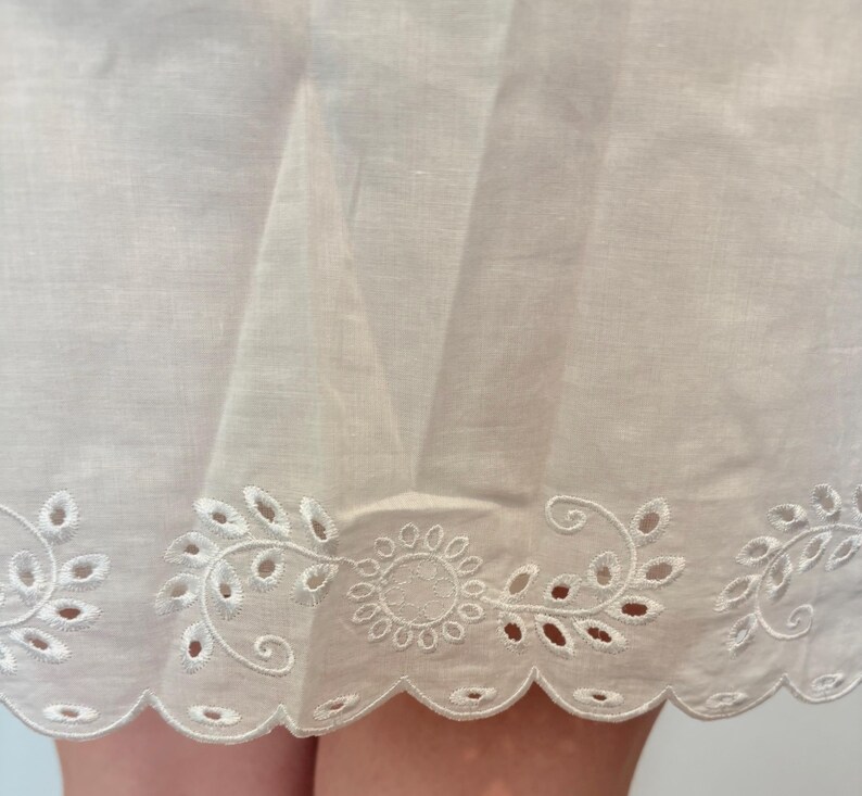 May include: Close-up of a white fabric with a scalloped edge. The edge features an embroidered floral design with cut-out details. The fabric appears to be lightweight and slightly translucent, suggesting a delicate texture.