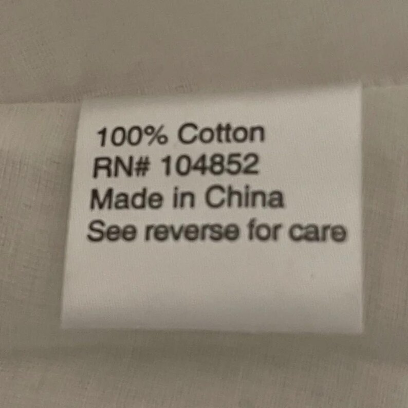 May include: A white rectangular tag with black text. The text reads "100% Cotton", "RN# 104852", "Made in China", and "See reverse for care". The tag is likely a clothing label.
