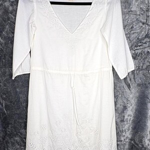 May include: White, knee-length dress with a V-neck and three-quarter sleeves. The dress features embroidered floral detailing along the neckline and hem, and a drawstring waist. The dress is hanging on a black hanger.