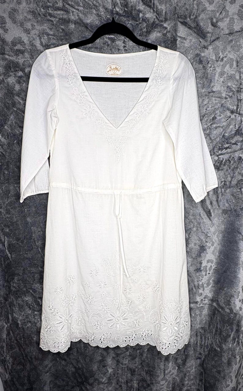 May include: White, knee-length dress with a V-neck and three-quarter sleeves. The dress features embroidered floral detailing along the neckline and hem, and a drawstring waist. The dress is hanging on a black hanger.