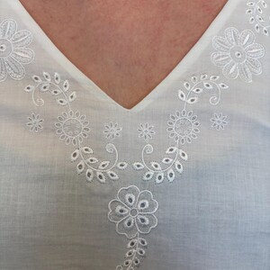May include: White V-neck top with intricate white floral embroidery around the neckline. The embroidery features detailed flower and leaf patterns. The fabric appears to be lightweight and breathable, perfect for warm weather.