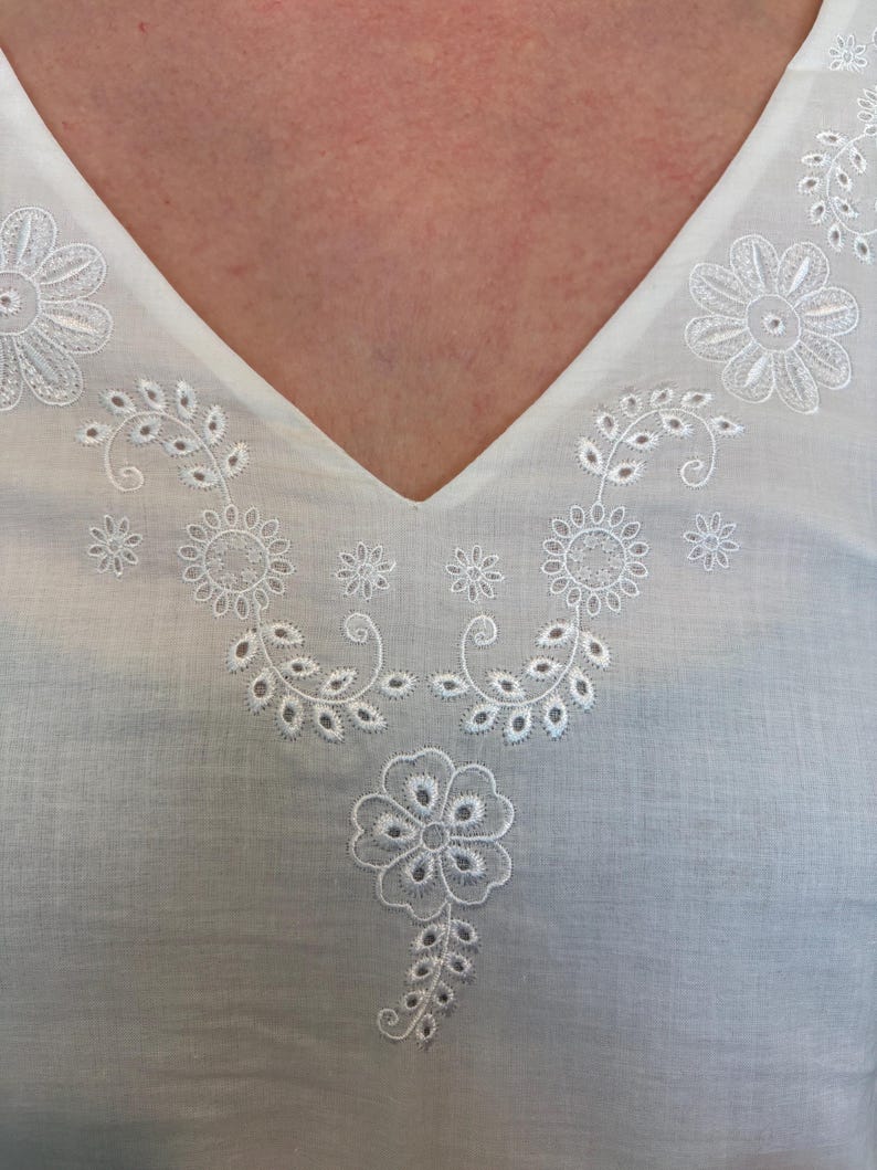 May include: White V-neck top with intricate white floral embroidery around the neckline. The embroidery features detailed flower and leaf patterns. The fabric appears to be lightweight and breathable, perfect for warm weather.
