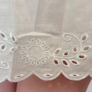 May include: Close-up of white fabric with intricate eyelet embroidery. The design features a floral motif with a scalloped edge. The fabric appears to be lightweight and delicate, suggesting a garment or home decor item.