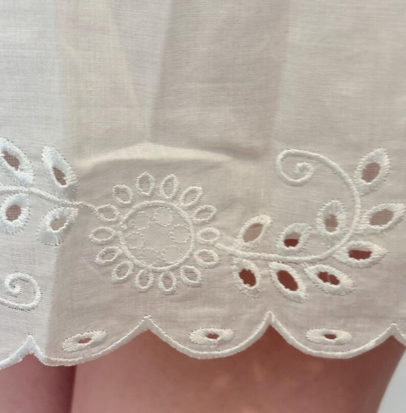 May include: Close-up of white fabric with intricate eyelet embroidery. The design features a floral motif with a scalloped edge. The fabric appears to be lightweight and delicate, suggesting a garment or home decor item.