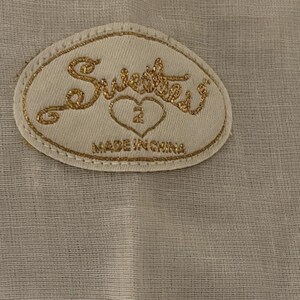 May include: A beige fabric tag with gold embroidered text and a gold border. The tag reads "Sweetest" above a heart with the number "2" inside. Below the heart, the tag reads "MADE IN CHINA".