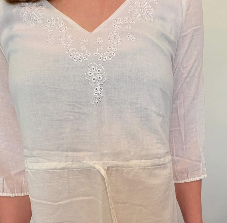 May include: White, lightweight tunic with a V-neck and three-quarter sleeves. The tunic features delicate white floral embroidery around the neckline and a drawstring waist. The fabric appears to be a soft, breathable material, suitable for warm weather.