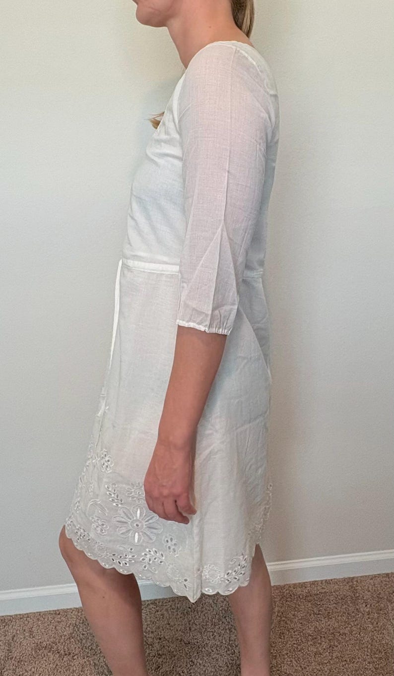 May include: White eyelet dress with three-quarter sleeves and a tie waist. The dress features a floral embroidered hemline. The fabric appears to be lightweight and suitable for warm weather. The dress is knee-length.