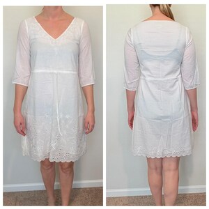 May include: White, knee-length dress with a V-neck and three-quarter sleeves. The dress features embroidered detailing around the neckline, waist, and hem. The fabric appears lightweight and semi-sheer, suitable for warm weather.