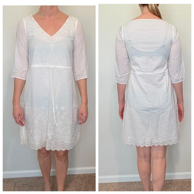 May include: White, knee-length dress with a V-neck and three-quarter sleeves. The dress features embroidered detailing around the neckline, waist, and hem. The fabric appears lightweight and semi-sheer, suitable for warm weather.