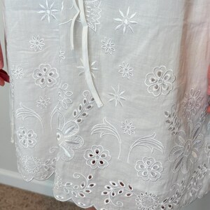 May include: White eyelet fabric with floral and leaf embroidery. The fabric features a scalloped edge and a drawstring. The embroidery includes various flower and leaf designs, creating a delicate and detailed pattern. The overall design is light and airy.