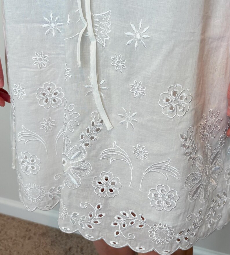 May include: White eyelet fabric with floral and leaf embroidery. The fabric features a scalloped edge and a drawstring. The embroidery includes various flower and leaf designs, creating a delicate and detailed pattern. The overall design is light and airy.
