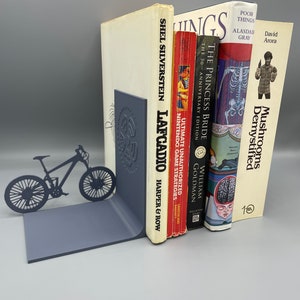 May include: A set of bookends featuring a black metal silhouette of a mountain bike. The bookends are holding up a stack of books, including "The Princess Bride", "Poor Things", "Mushrooms Demystified", "Ultimate Unauthorized Nintendo Game Strategies", and "Lafcadio" by Shel Silverstein.