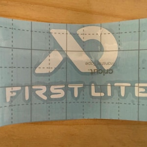 May include: A light blue Cricut transfer tape sheet with a white logo and the words "FIRST LITE". The logo is a stylized letter "X" and a "D" shape. The sheet is on a wooden surface.
