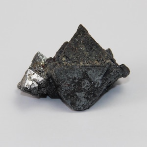 May include: A dark grey, metallic, octahedral crystal with a rough, textured surface.