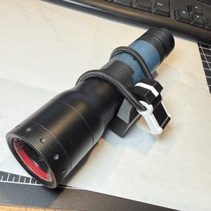 May include: A black cylindrical flashlight with a blue textured grip section. A black cord is wrapped around the body, secured by a small black and white plastic clip mechanism. The flashlight features a circular lens with a red inner ring.