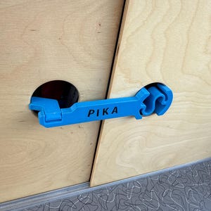 May include: A light blue handle with the word "PIKA" printed on it is attached to a wooden surface. The handle is inserted into a black hole. The wooden surface is light brown and has a diagonal seam.