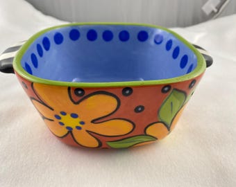 Può includere: Una colorata ciotola in ceramica di forma quadrata con due manici. L'interno è blu, il bordo è verde e l'esterno è decorato con fiori arancioni e foglie verdi. I manici sono a righe bianche e nere.