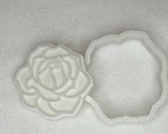 Può includere: Un tagliabiscotti a forma di rosa in plastica bianca e il suo contorno abbinato. Il tagliabiscotti è un design floreale dettagliato, perfetto per la cottura. Il contorno ha una forma semplice e arrotondata. Gli oggetti sono su una superficie bianca.