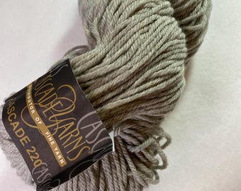 Può includere: Un gomitolo di filato Cascade 220 in un colore grigio tenue. Il filato è ben avvolto e ha un aspetto strutturato. L'etichetta recita "Cascade Yarns, Distributor of Fine Yarn".