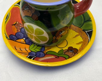 Può includere: Un set di tazza e piattino in ceramica dipinti a mano. La tazza ha una base verde con disegni di scimmie e frutta, un manico rosso e un bordo giallo con pois neri. Il piattino è giallo con illustrazioni di frutta e scimmie.