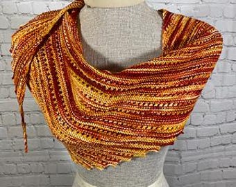 Può includere: Uno scialle lavorato a maglia nei toni dell'arancione, giallo e rosso, drappeggiato su un manichino. Lo scialle presenta un motivo a righe e un bordo smerlato. Il filato sembra essere una miscela di colori, creando un aspetto caldo e strutturato.