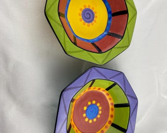 Può includere: Due ciotole decorative ottagonali con vivaci disegni astratti. Una ciotola presenta un bordo verde, centro rosso e giallo e dettagli blu. L'altra ciotola ha un bordo viola, centro verde e marrone e dettagli blu. Entrambe le ciotole hanno linee nere.