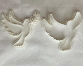 Può includere: Due stampini per biscotti in plastica bianca a forma di colombe, uno con un ramo di ulivo nel becco.