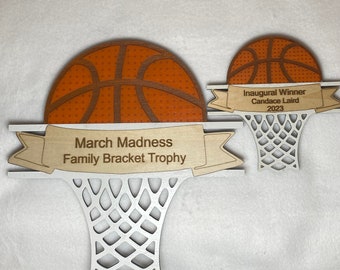 Può includere: Un trofeo a forma di canestro da basket in legno con una palla da basket marrone e uno striscione che recita "March Madness Family Bracket Trophy". Un trofeo a forma di canestro da basket in legno più piccolo con una palla da basket marrone e uno striscione che recita "Inaugural Winner Candace Laird 2023".