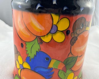 Può includere: Un vaso in ceramica dal design vivace. Il vaso presenta uno sfondo rosso con zucche arancioni, fiori gialli e un tucano blu. Il bordo è nero con accenti bianchi.