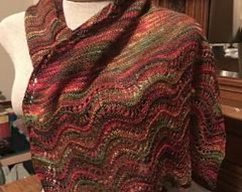 Può includere: Uno scialle lavorato a mano a maglia nei toni del rosso, marrone e verde, esposto su un manichino. Lo scialle presenta un motivo ondulato e strutturato con un bordo smerlato. Il filato sembra avere un effetto cromatico variegato, creando un aspetto ricco e stratificato.