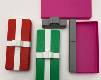 Può includere: Una collezione di scatole regalo e componenti colorati. Le scatole sono rosse, verdi e rosa, con nastri bianchi. I componenti includono un fiocco grigio e un coperchio rosa. Gli articoli sono disposti su una superficie bianca.