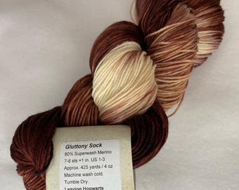 Può includere: Un gomitolo di filato marrone, beige e crema con un'etichetta che recita "Gluttony Sock 80% Superwash Merino 7-8 sts = 1 in. US 1-3 Approx. 425 yards / 4 oz Machine wash cold. Tumble Dry. Leaving Hogwarts"