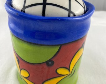 Può includere: Un barattolo di ceramica con coperchio. Il barattolo è cilindrico con un bordo blu e una base rossa decorata con disegni floreali verdi e gialli. Il coperchio è bianco con un motivo a griglia nera e un pomello blu.