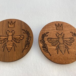 May include: Two round wooden coasters with a laser-etched design of a bee wearing a crown and surrounded by leaves. The coasters are a dark brown color.