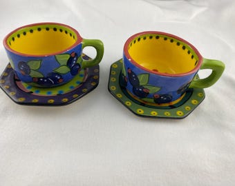 Può includere: Due tazze da tè e piattini in ceramica. Le tazze hanno interno giallo, esterno blu con un disegno di bacche e manici verdi. I piattini esagonali hanno bordi verdi e viola e un motivo a pois gialli.