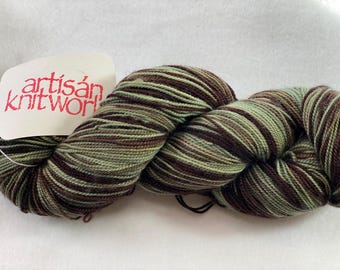Può includere: Un gomitolo di filato a righe marroni, verdi e bianche. Il filato è etichettato "artisan knitwork".