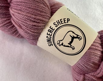 Può includere: Un gomitolo di filato color rosa polvere con un'etichetta bianca che dice "Sincere Sheep" e ha un'illustrazione in bianco e nero di una pecora.
