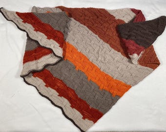 Può includere: Uno scialle lavorato a maglia con un motivo a righe diagonali. Lo scialle presenta strisce alternate arancioni, marroni, grigie e beige. I bordi sono smerlati e rifiniti con un bordo marrone scuro. Lo scialle è piegato su una superficie bianca.