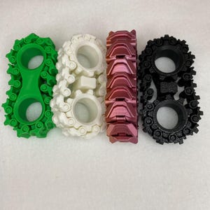May include: Four 3D printed track sets in different colors: green, white, pink, and black. Each track set is made of interlocking pieces that form a continuous loop.