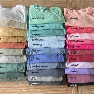 May include: A stack of folded t-shirts in various colors, including white, ivory, butter, mustard, yam, sandstone, bay, light green, blue spruce, moss, island reef, chalky mint, seafoam, hydrangea, chambray, blue jean, flo blue, washed denim, denim, gray, pepper, peachy, crunch berry, blossom, watermelon, heliconia, brick, chili, orchid, violet, berry, and black.