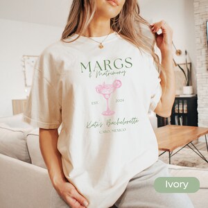 May include: Ivory t-shirt with a green and pink graphic design. The graphic features a margarita glass with the text "MARGS & Matrimony Est. 2024 Kate's Bachelorette Cabo, Mexico".