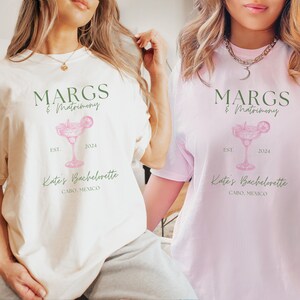 May include: Two white t-shirts with a green and pink design. The design features a margarita glass with a lime wedge, the text "MARGS & Matrimony", "EST. 2024", and "Kate's Bachelorette CABO, MEXICO".