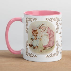May include: White ceramic mug with a pink interior and handle. The mug features a vintage illustration of a cat and a woman in a pink dress. The illustration is framed with a brown floral border. The mug is placed on a wooden surface.