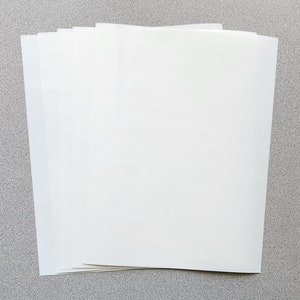 May include: A stack of white, rectangular sheets of paper, slightly fanned out. The paper has a smooth, clean surface and appears to be unused. The sheets are arranged on a gray surface.