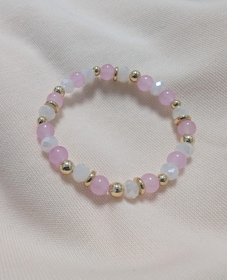 Puede incluir: Una pulsera el&aacute;stica que presenta una alternancia de cuentas rosas lisas, cuentas blancas facetadas y peque&ntilde;as cuentas espaciadoras doradas dispuestas en un patr&oacute;n circular. El dise&ntilde;o consiste en una secuencia repetitiva de estos tres tipos.